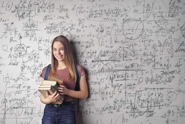 A young woman holds books and behind her is a whiteboard filled with mathematical equations